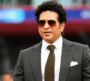 Sachin Tendulkar in a formal black suit and sunglasses at a cricket stadium during a public appearance.