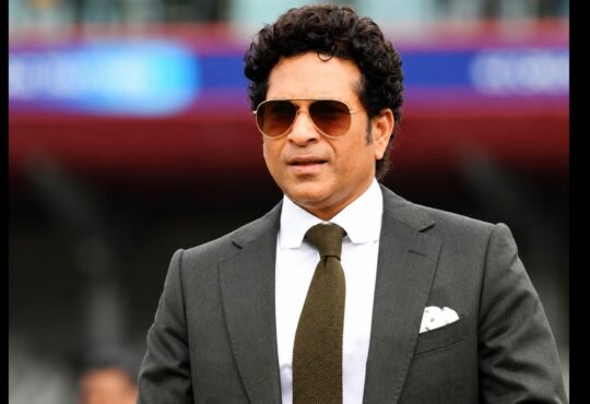 Sachin Tendulkar in a formal black suit and sunglasses at a cricket stadium during a public appearance.